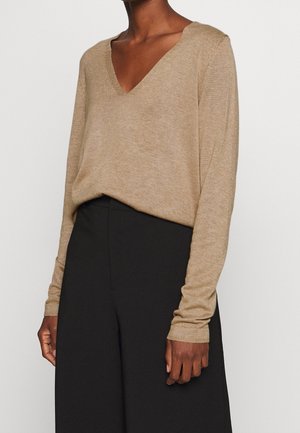 Beige, lichte gebreide trui met een V-hals en lange mouwen, met een relaxed fit en geribbelde manchetten. Ggedragen met zwarte broeken.