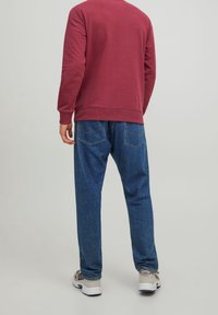 Felpa bordeaux con polsini e orlo a coste, abbinata a jeans in denim blu con tasche posteriori e vestibilità comoda. Scarpe da ginnastica grigie e bianche.