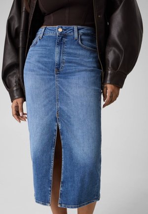 Femme portant une jupe midi en denim bleu avec une fente avant et une taille haute, associée à un top noir et une veste en cuir marron foncé.