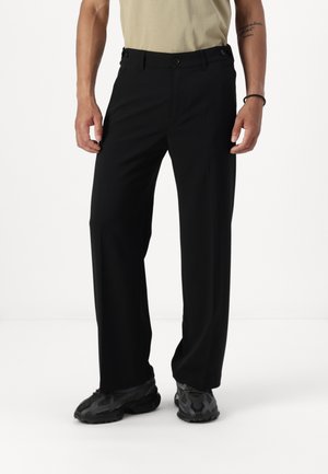 Man die losse zwarte broek, zwarte sneakers en een beige T-shirt draagt, staat met ontspannen handen langs het lichaam tegen een effen achtergrond.