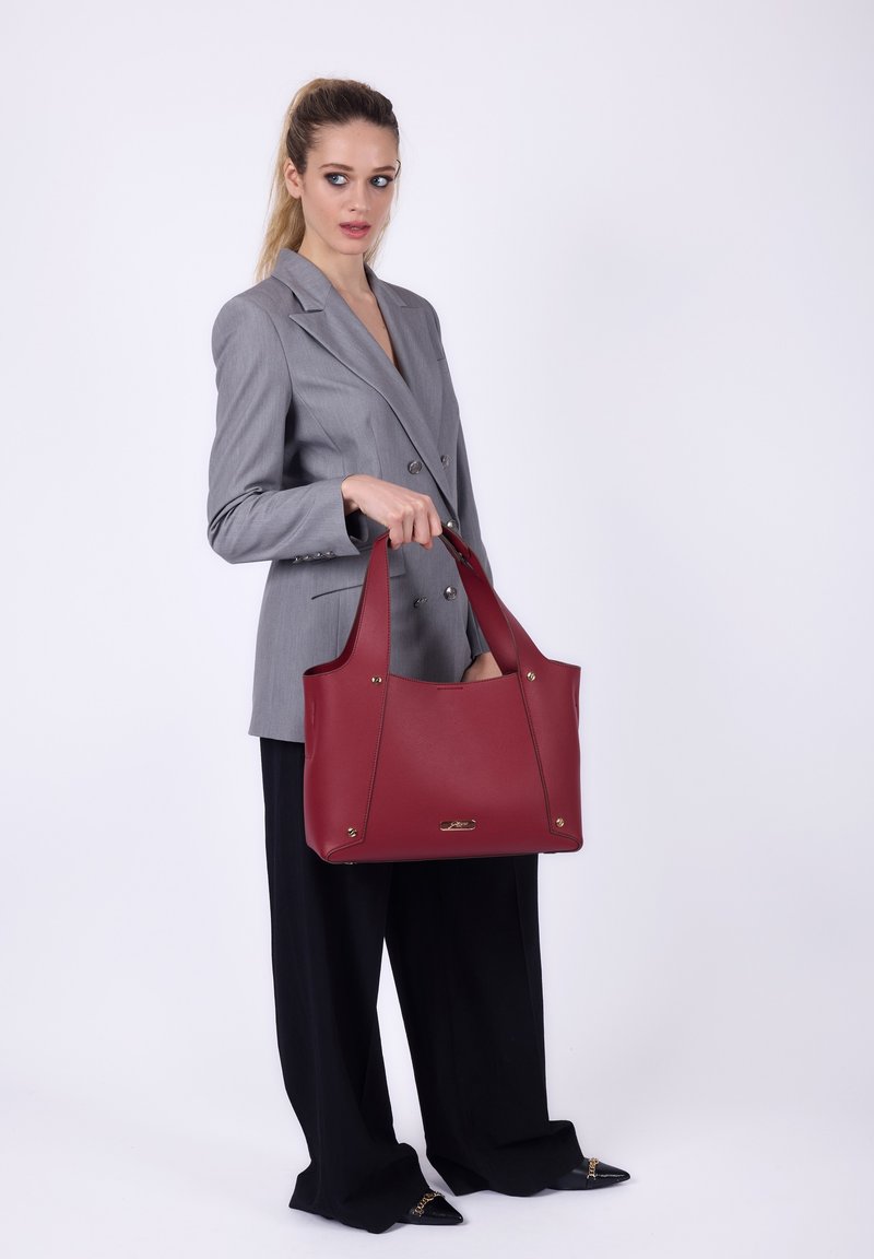 Vrouw in een grijze blazer en zwarte wijde broek, met een grote bordeauxleren tote bag tegen een witte achtergrond.