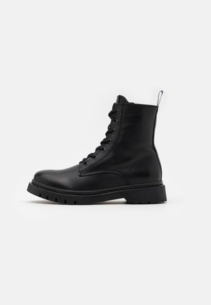 Bottine noire en cuir à lacets avec semelle épaisse et languette à l'arrière, vue de profil sur fond blanc.