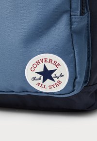 Blå lerret ryggsekk med en hvit, sirkulær lapp med en marineblå stjerne og rød tekst "CONVERSE ALL STAR" og "Chuck Taylor." Strukturerte tekstiler.