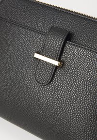 Portefeuille en cuir noir texturé avec un fermoir métallique horizontal en ton or. Dispose d'une fermeture éclair et de coutures visibles pour la durabilité.
