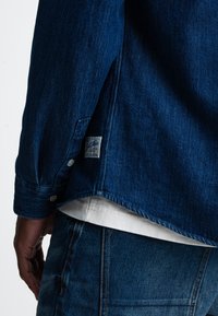 Donkerblauwe denimshirt met een puntkraag, knoopsluiting bij de manchetten en een label aan de linkerkant nabij de zoom. Witte onderhemd zichtbaar.