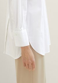 Chemise en coton blanc à manches longues, avec des poignets boutonnés, une silhouette droite et un ourlet incurvé, associée à un pantalon beige clair.