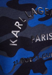 Textilie s maskáčovým vzorem v odstínech modré a tmavě šedé, s vyvýšeným nápisem: "KARL LAGERFELD PARIS" a "21 RUE ST-GUILLAUME."