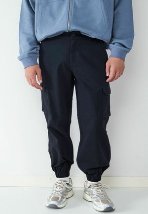 Person wearing black cargo jogger pants and white sneakers with gray accents, standing against a plain white background.