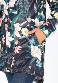 Impermeabile floreale con sfondo blu navy, caratterizzato da vivaci fiori rosa e bianchi. Dotato di tasche laterali e chiusure a scatto sui polsini.