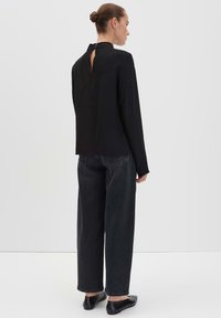 Donna con i capelli raccolti a chignon, che indossa un top nero a maniche lunghe con un'apertura a chiave sulla schiena e jeans larghi scuri, in piedi su uno sfondo neutro.