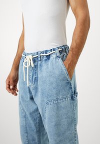 Világoskék farmer jogger nadrág, elasztikus derékkal, állítható zsinórral, oldalsó zsebekkel és laza szabással. Megviselt textúra részlet.