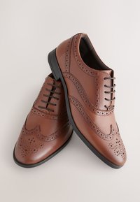 Paire de chaussures habillées brogues en cuir marron avec perforations décoratives et semelles noires, disposées en croix sur un fond clair.