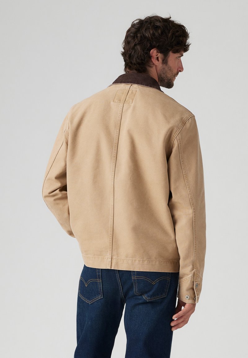 Chaqueta color beige con un cuello marrón, con un diseño simple, tela suave y puños con botones; mostrada desde la parte posterior con jeans azul oscuro.