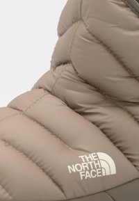 Botte isolante beige avec un design matelassé, une texture lisse et un logo. La tige présente des coutures visibles et une semelle en caoutchouc contrastante.
