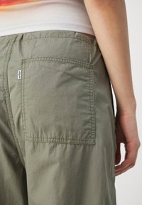 As calças verde oliva apresentam um grande bolso traseiro com costuras combinadas. O tecido aparenta ser leve com uma textura suave. Etiqueta do logotipo visível.