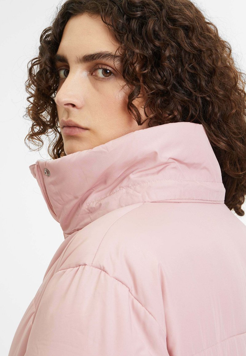 Gezwollen roze jasje met een hoge kraag, met een gladde textuur en zichtbare stiksels. Profielfoto van het model in zacht licht.