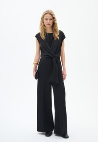 Jumpsuit noir avec un haut drapé et un détail noué à la taille, présentant des jambes larges. Le mannequin tient une pochette noire et porte un bracelet en or.