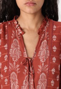 Top couleur rouille avec un col à volants, détail à nouer et motifs floraux blancs. Présente un tissu texturé avec des accents tissés le long des bords.