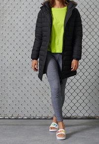 Svart pufferjacka med huva, ljusgrön T-shirt, grå leggings och vita slides med färgglad text. Grå kedje-länkbakgrund.