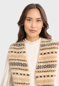 Fleece-Weste mit einem beigen und gemusterten Design, das schwarze und pinke Akzente aufweist, versehen mit einer Knopfleiste und weicher Textur.