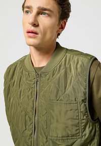 Gilet verde oliva trapuntato con zip, colletto rotondo e tasca laterale; presenta una texture liscia e lucida e un motivo a rombi.