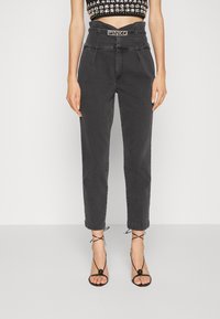 Pantalones de mezclilla negros con cintura alta, frente plisado y piernas ajustadas. Presenta un cinturón a juego con hebilla de logo. Se llevan con sandalias negras de tiras.