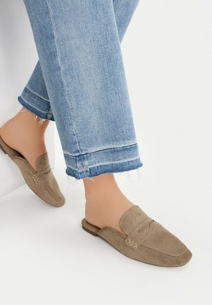 Füße in hellblauen verkürzten Jeans und beigen, wildledernen, schuhlosen Loafers mit niedrigen Absätzen vor weißem Hintergrund.