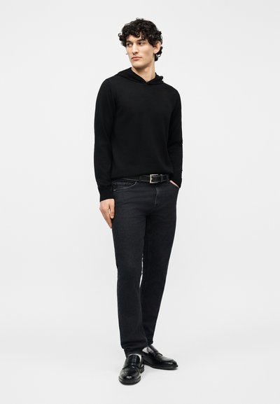 Νέος με κατσαρά μαλλιά, φορούσε μαύρο φούτερ με κουκούλα, σκούρα τζιν, μαύρη ζώνη και μαύρα επίσημα παπούτσια, στέκεται με το ένα χέρι στην τσέπη.