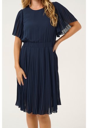 Frau trägt ein marineblaues, plissiertes Midikleid mit transparenten, flatternden Ärmeln und tailliertem Bund, steht mit einer Hand in der Hüfte.