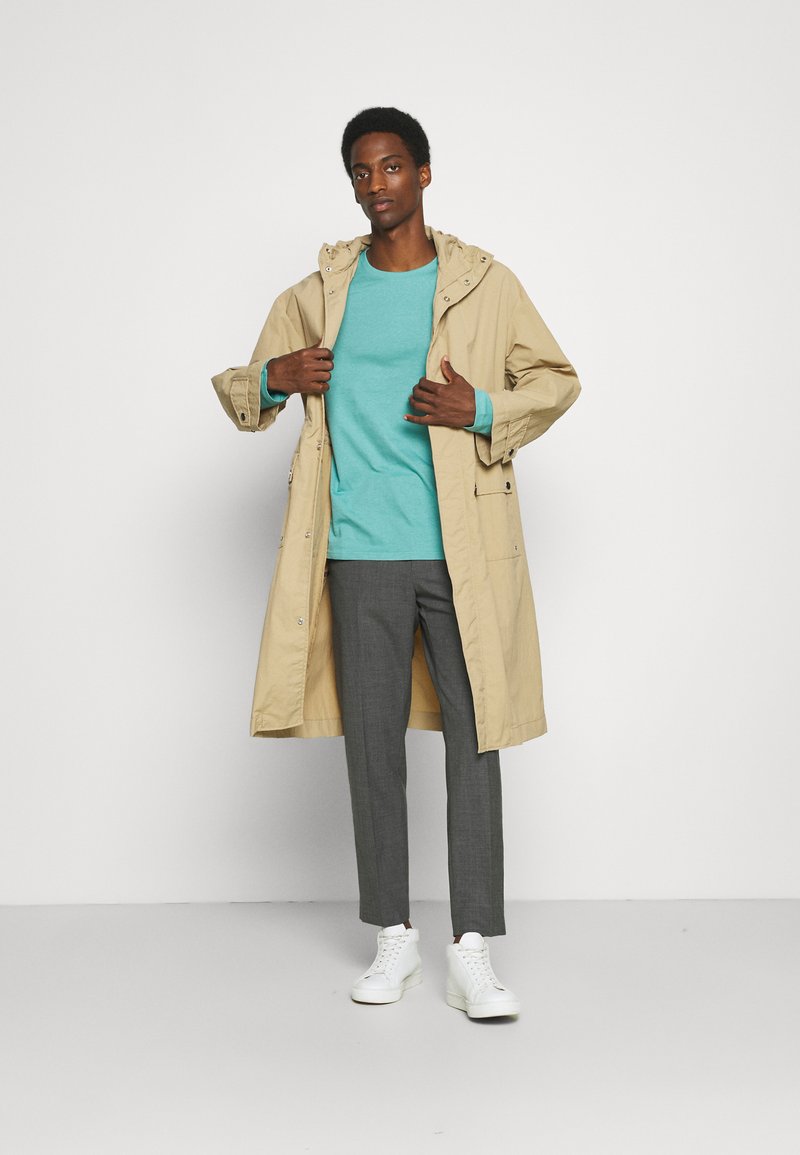 Beige gewatteerde jas over een teal sweatshirt, grijze broek en witte sneakers. De jas heeft een ritssluiting en knoopsluiting met zijzakken.