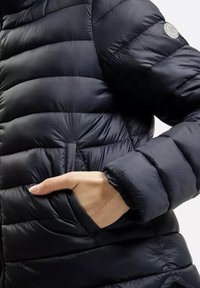 Czarna kurtka puchowa z pikowanym wzorem, teksturowanym materiałem i kieszenią po boku. Zauważalny okrągły logo na lewym ramieniu.