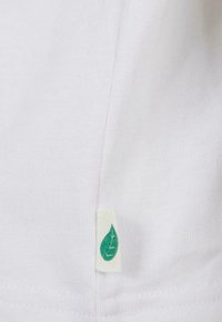 Hvit tekstilmerke med design av et grønt blad, detaljert med en linjeillustrasjon, sydd til kanten av plagget.