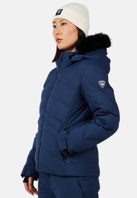 Giacca a vento blu navy con chiusura a zip, cuciture orizzontali e cappuccio sfoderato in pelliccia staccabile. Abbinata a un berretto in maglia crema.