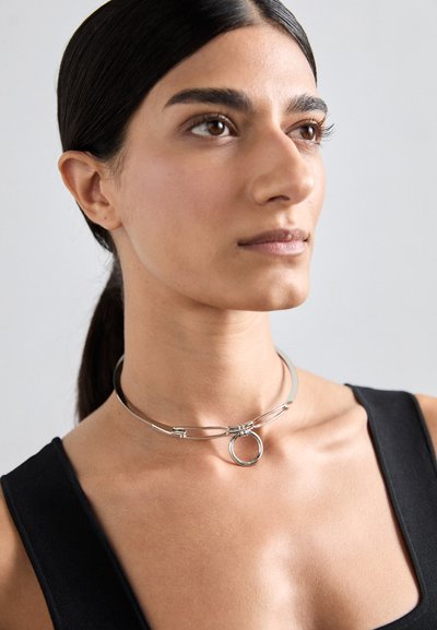 Choker en argent avec un pendentif circulaire, design épuré avec une surface lisse et des accents métalliques courbés, porté avec un top noir.
