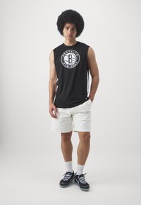 Camiseta sin mangas negra con logo blanco de Brooklyn, pantalones cortos blancos, zapatillas negras con acentos blancos, de pie contra un fondo claro.