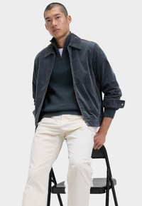 Herrencordjacke in Dunkelgrau mit kragenförmigem Ausschnitt, kombiniert mit einem dunkelgrünen Pullover und weißen Hosen. Der strukturierte Stoff betont das Design.