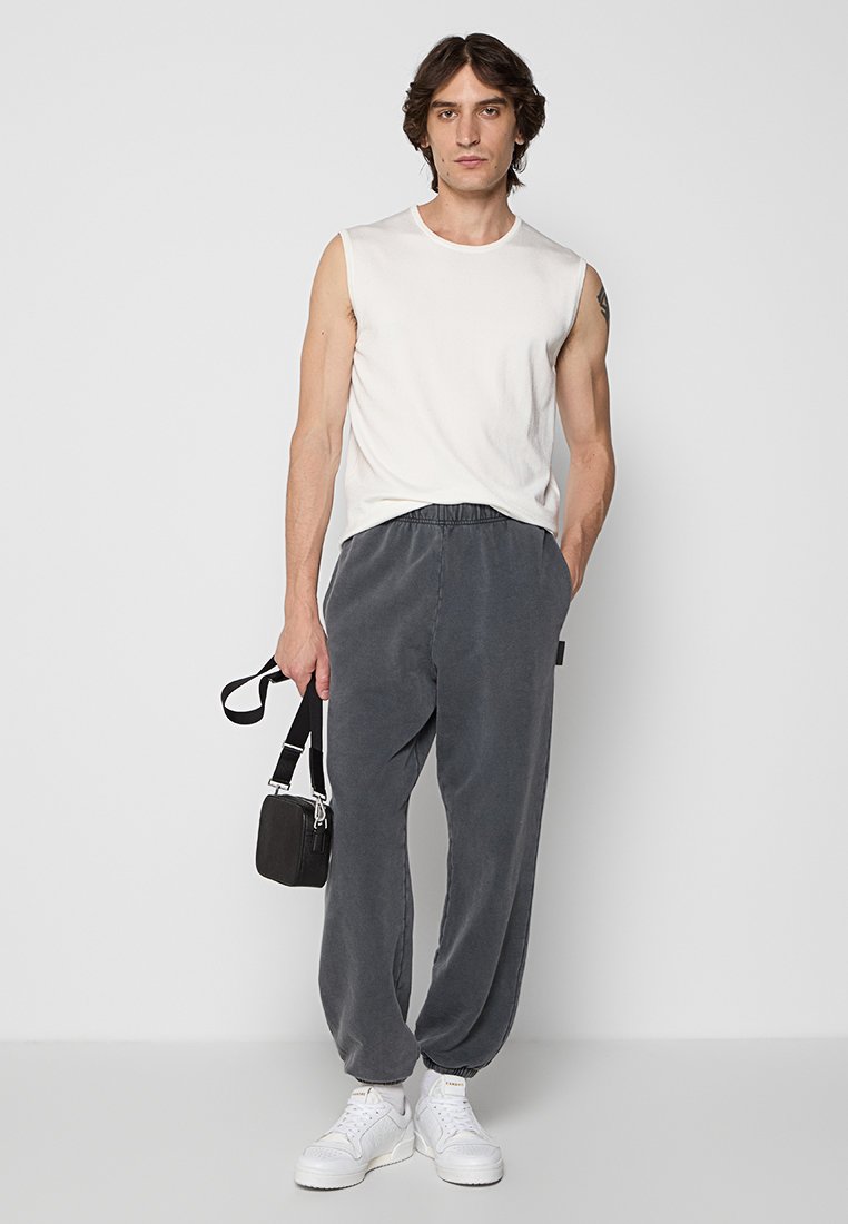 Top sin mangas de color crema para hombre con pantalones de cintura elástica gris, tejido texturizado y bolsa cruzada negra. Zapatillas blancas completan el look.