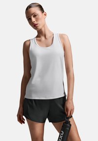 Hvid atletisk tanktop med rund halsudskæring og tætsiddende pasform, kombineret med sorte shorts. Modellen holder en sort vandflaske.