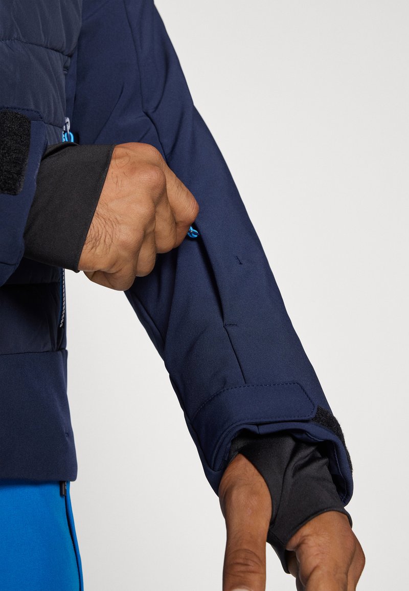 Marineblauwe geïsoleerde jas met een ritszak op de arm, voorzien van een zwarte binnenvoering, klittenbandsluiting en verstelbaar elastisch koord.