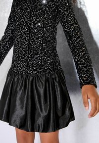 Rochie neagră cu un top cu mărgele și mâneci lungi, având o fustă subțire din satin negru lucios. Textură netedă și cu un corset ajustat.