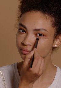 Application de correcteur en cours à l'aide d'un petit pinceau. Teint de peau moyen avec une texture lisse. Mise au point sur un œil, avec un produit de couleur claire appliqué.