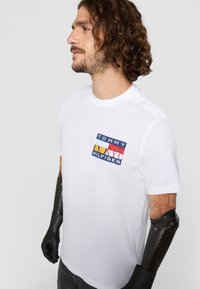 Hvid t-shirt med korte ærmer, der har et farverigt rektangulært "TOMMY HILFIGER" logo-patch på venstre bryst; glat stofstruktur.