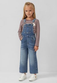 Denim overall met wijde pijpen, vervaagde blauwe kleur en voorvak, gecombineerd met een gestreepte long-sleeve top. Witte sneakers maken de look compleet.