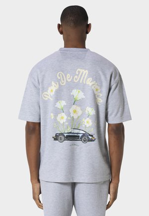 Mann iført lysegrå skjorte og bukser, på baksiden av skjorten vises grafikk av en svart bil med hvite blomster og teksten "Pas De Monaco" over.