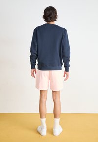 Sudadera de cuello redondo en color marino con puños de canalé, combinada con pantalones cortos rosa claro. El modelo lleva zapatillas blancas y calcetas hasta el tobillo, visto desde atrás.