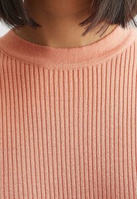 Geribbelde gebreide trui in licht koraal, met een ronde halslijn en een zachte textuur. De stof vertoont een consistent verticaal patroon.