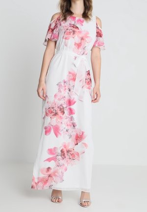 Femme portant une robe maxi blanche avec des imprimés floraux roses, des manches épaules dénudées et une ceinture à la taille, associée à des talons argentés ouverts à l'avant.