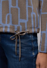 Top de manga larga con estampado marrón y azul, con cintura ajustable mediante cordón, combinado con vaqueros de mezclilla azul oscuro con un ligero detalle desgastado.