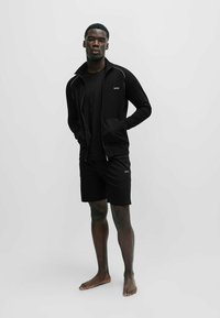 Svart treningsdrakt som inkluderer en jakke med glidelås og sidelommer, i kombinasjon med shorts. Begge deler har et merke med logo.