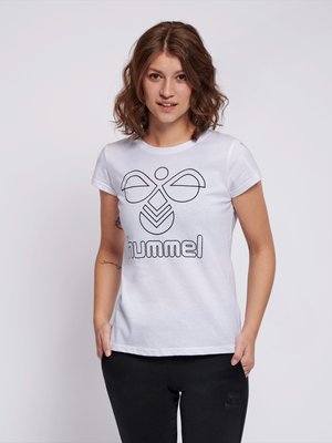 Kvinde med bølget brunt hår iført en hvid t-shirt med sort hummel-logo og sorte bukser, stående med hænderne i lommerne.